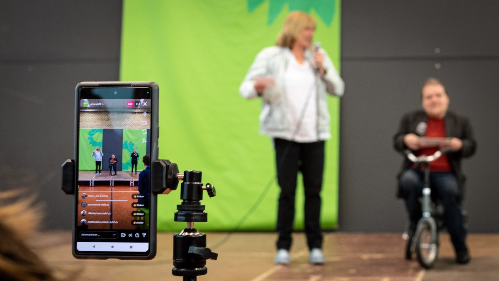 Im Vordergrund ein Handy auf Stativ, welches im Hintergrund zwei Menschen auf der Bühne filmt.
