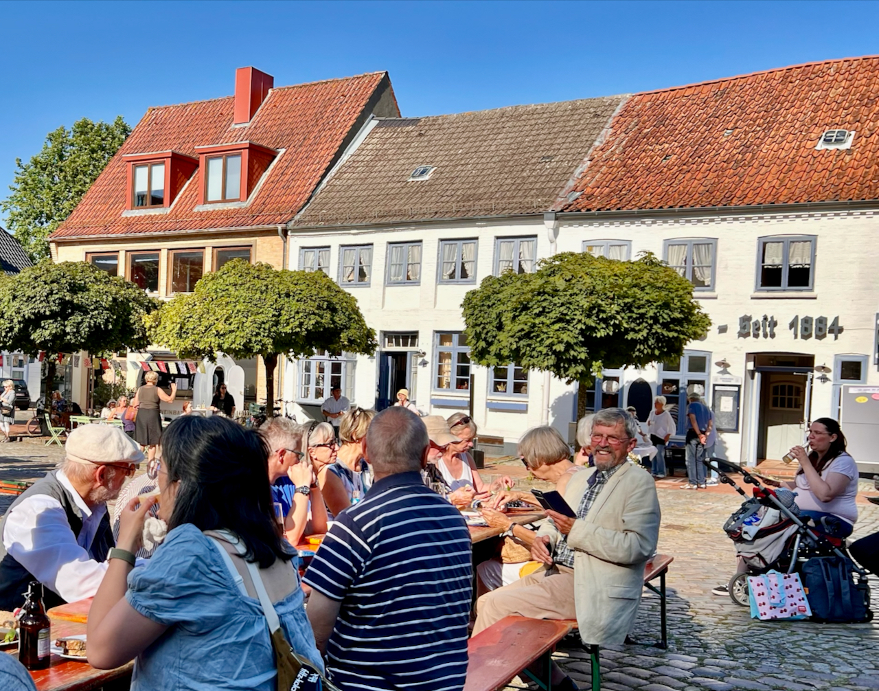 Foto mit Menschen, die an einem Tisch auf einem Platz in der Sonne sitzen.