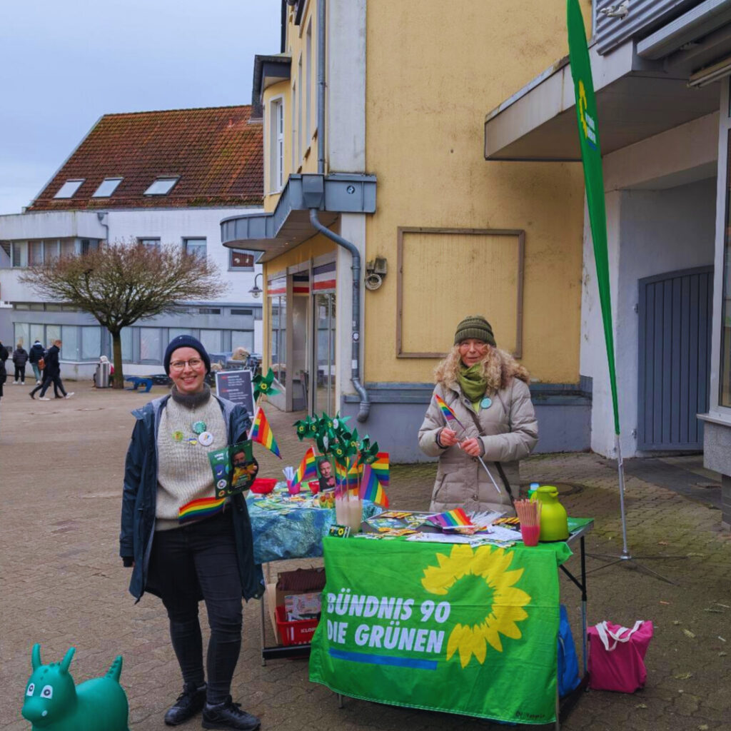 Meike und Elke am Grünen Wahlkampftisch. Die Beachflag ist zu sehen. Meike hält Flyer und eine Regenbogenflagge in ihren Händen.
